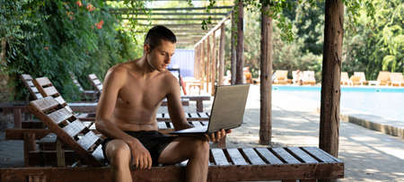 caucasian male businessman with a computer sits on a wooden deck chair in the shade near the pool and works remotely. freelancer concept in hotel. copyspace.の写真素材