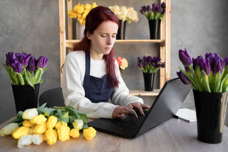 Portrait of a florist in an apron working in her own flower shop, using a laptop to organize logistics and delivery, taking orders online.の写真素材