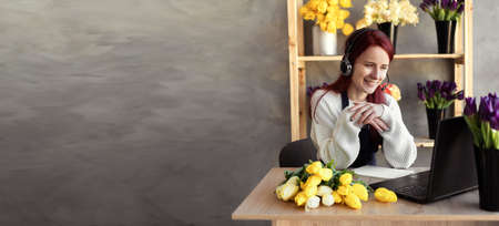 Young beautiful female florist in blue apron business owner using headset and computer while talking to customers online taking order online. Banner. copyspace.の写真素材