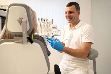 man in a smock doctor dentist shows an artificial plastic jaw bite for a female patient sitting in the dentists office in the office near the dentist. Medical treatment of enamel. orthodontist workingの写真素材