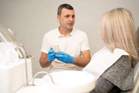 a male orthodontist conducts a consultation on dental prosthetics. Dentist doctor shows an artificial plastic jaw with dental implants. Background of modern clinic.の写真素材