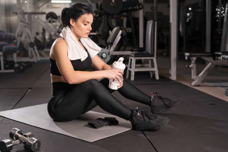 Young tired female athlete resting in the gym, sportswoman sits on a fitness mat with water after training, healthy lifestyleの写真素材