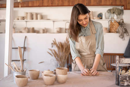 Young beautiful woman with long hair in an apron creates a handmade ceramic bowl from clay. Creative workshop. The concept of skill and entrepreneurship.の写真素材