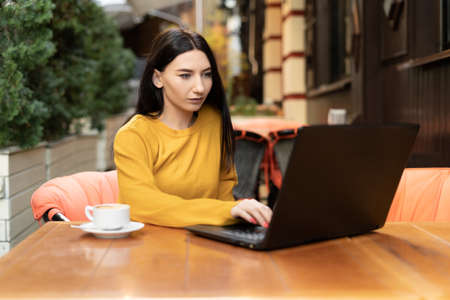 beautiful girl is sitting outdoors in a cafe in an orange sweater looking at a laptop. A young brunette woman works remotely, shopping online. warm autumn day.の写真素材