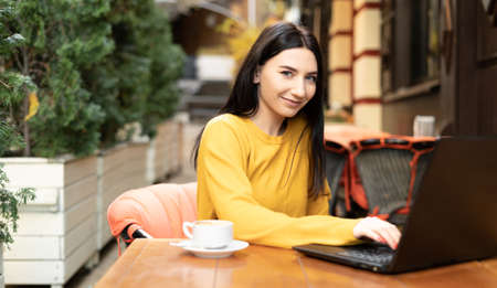 beautiful smiling girl is sitting outdoors in a cafe in an orange sweater looking at the camera. A young brunette woman works remotely, shopping online. warm autumn day.の写真素材