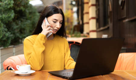 Corporate communication. beautiful businesswoman talking on mobile phone while working on laptop remotely in outdoor autumn cafe.の写真素材