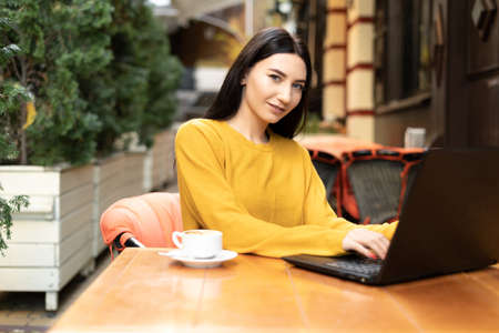 beautiful smiling girl is sitting outdoors in a cafe in an orange sweater looking at the camera. A young brunette woman works remotely, shopping online. warm autumn day.の写真素材