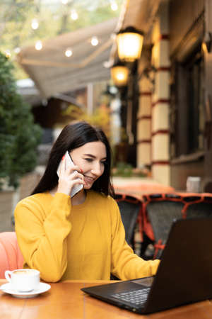 Corporate communication. beautiful businesswoman talking on mobile phone while working on laptop remotely in outdoor autumn cafe.の写真素材