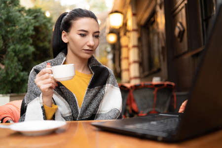 A girl in a cafe on the street wrapped in a plaid, enjoying a sunny warm autumn day. Cup of coffee tea in hands Young brunette working behind a laptop.の写真素材
