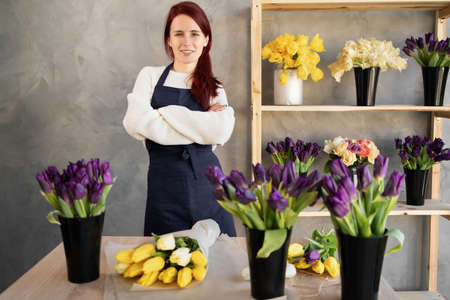 Young caucasian sales girl with crossed arms in a work apron smiles happily while standing at a window display in a flower shop.の写真素材