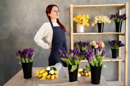 Young caucasian saleswoman girl standing and suffering from back pain while working, standing by the window display in a flower shop.の写真素材