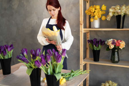 Portrait of a smiling florist owner standing in an apron with a bouquet of yellow tulips near a shop window surrounded by plants.の写真素材