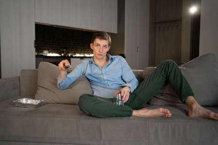 Caucasian man in casual clothes sits at home in the evening on the sofa with a TV remote control and switches channels.の写真素材
