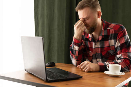 caucasian bearded businessman works overtime and feels tired eyes in the office, massaging the bridge of the nose eye pain, vision problemsの写真素材