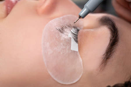 Eyelash extension process. Close-up portrait of a young girl, a woman with eyelashes, closed eyes and a beautician's hand making markings with a pencil on a patch.の写真素材