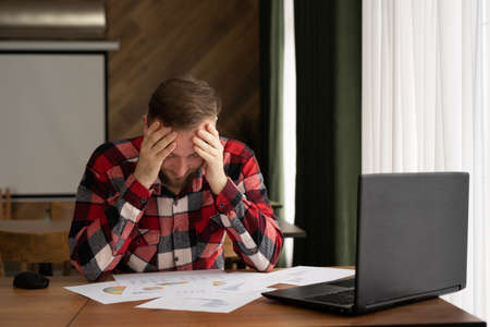 Portrait of a tired nervous bearded businessman in casual clothes working in a cafe, sitting with a laptop, problems of a remote employee, a man in the stress of a failed projectの写真素材