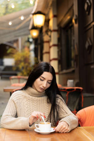 young beautiful caucasian woman sits in a cafe in a sweater stirs sugar in coffee. copy spaceの写真素材