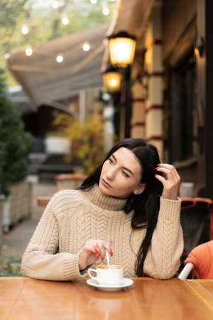 young beautiful caucasian woman sits in a cafe in a sweater stirs sugar in coffee. copy spaceの写真素材