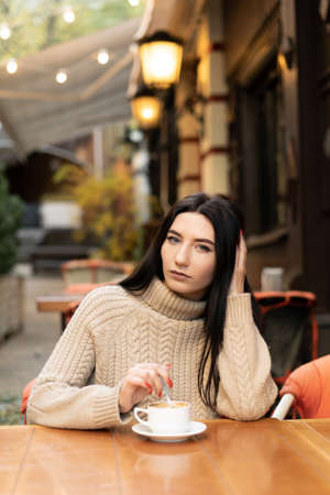 Portrait of a joyful young woman enjoying a cup of coffee in a cafe. Smiling beautiful girl drinking hot drink in autumn. Excited lady in a sweater on an autumn day.の写真素材