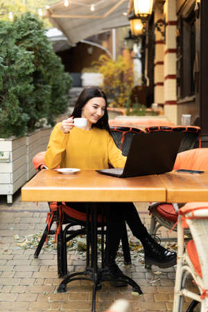 Portrait of a young beautiful businesswoman enjoying coffee while working on a laptop computer in a cafe outdoors in autumn, during a morning breakfastの写真素材