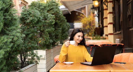 Portrait of a young beautiful businesswoman enjoying coffee while working on a laptop computer in a cafe on the street in autumn, during the morning breakfast. Banner, copy space.の写真素材