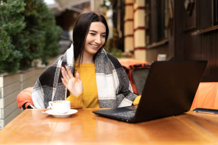 A woman sits with a laptop and a cup of coffee on the terrace in a cafe in autumn. Bright orange sweater and plain. Freelancer remote work concept. copyspace.の写真素材
