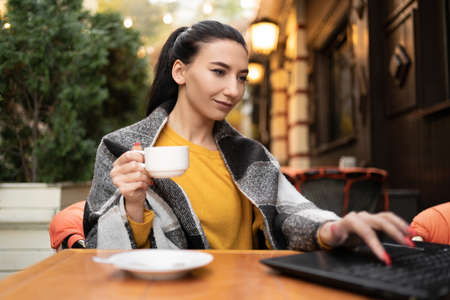 A woman sits with a laptop and a cup of coffee on the terrace in a cafe in autumn. Bright orange sweater and plain. Freelancer remote work concept. copyspace.の写真素材