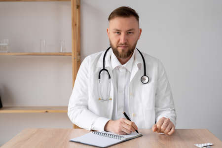 happy young male doctor wears white medical coat and stethoscope looking at camera. Smiling doctor, general practitioner consulting a patient online via video call. close-up headshot portraitの写真素材