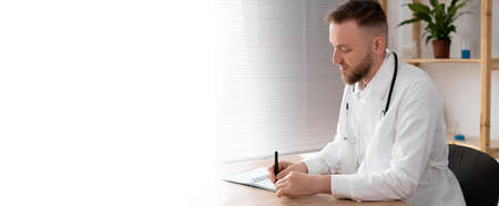 side view of a male doctor in the office in a white coat with a stethoscope working at the table. A serious doctor is waiting for a patient in a bright office ready to work. Banner. copyspace.の写真素材
