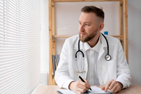 A caucasian doctor sits at a table in a bright office in a white coat with a stethoscope fills in the patient's hospital journal. Medicine, healthcare concept.の写真素材