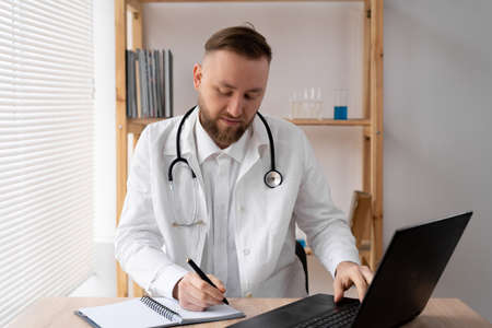serious male doctor using a laptop and writing notes in a medical journal while sitting at a table. in white coat and stethoscope working on computer at workplace.の写真素材