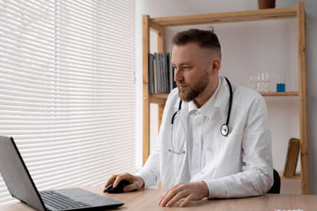 a male doctor in a white coat and with a stethoscope sits at a table in a hospital and types on a laptop.の写真素材