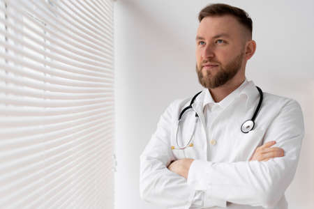 Portrait of a confident male doctor with crossed arms standing in a clinic near a window with jalousie. copyspace.の写真素材