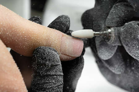 A manicurist applies an electric nail drill to a manicure on female fingers. A woman is getting a manicure of nails. Beautician removes old gel polish from client's nails.の写真素材