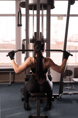 Shoulder lowering machine. Fitness woman is training in the gym. Upper body strength exercise for the upper back. back view without faceの写真素材