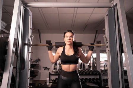 young adult girl doing barbell squats in the gym. A woman with a muscular body does lifting exercises.の写真素材
