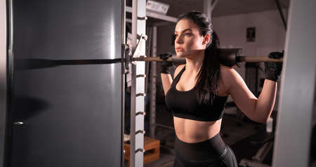 side view of a young adult girl doing barbell squats in the gym. A woman with a muscular body does lifting exercises.の写真素材