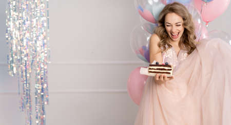 Millennial woman throwing a holiday party or celebration. a pretty woman in a pink dress holding a piece of cake in her hands stands on top of balloons with helium. Banner. copyspace.の写真素材