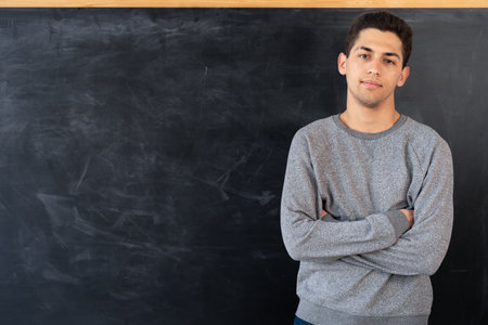 Happy young arab man standing with crossed arms on blackboard background, education concept, serious male teacher or arabic student, space for textの写真素材