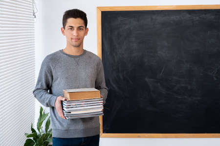 Portrait of arabic male teacher with books blackboard background. Back to school concept, education and knowledge, space for textの写真素材