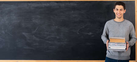 Young arabic teacher standing with books on blackboard background, university student with textbooks near chalk board in classroom, study and education concept, banner, space for textの写真素材