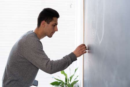 Young arab teacher leading lesson writes with chalk on blackboard, university student at chalk board, male arab 20s studying in class, side view tutor, education conceptの写真素材