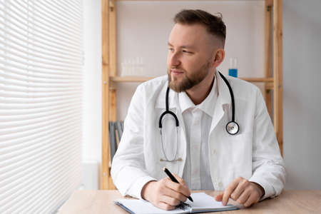 Portrait of a serious general practitioner in a white medical uniform sitting at a table in a hospital, a doctor or therapist with a stethoscope looks out window at work, medicine, healthcare conceptの写真素材