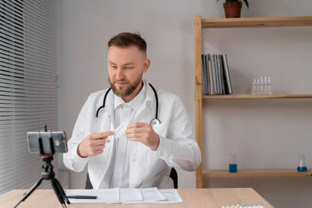Caucasian young male doctor in a bathrobe with a stethoscope makes an online consultation using a smartphone holds pills in his hands with his finger points to them. sale of medicinesの写真素材