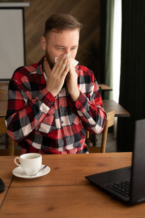 Ill caucasian man looking at laptop screen and sneezing nose in paper tissue, sitting on the office, empty space. Sinusitis illness, cold and influenza symptoms, allergy, air conditioner diseaseの写真素材