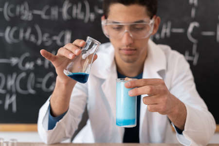 Male arabic chemistry teacher mixing liquids doing experience in college class, university student taking chemistry exam getting reaction of acids in glass flask against blackboard, science conceptの写真素材