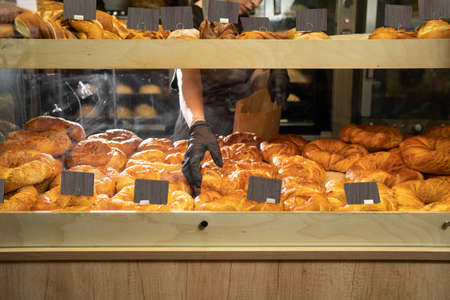 Freshly baked rolls behind a glass showcase of a store with price tags, seller's hand and delicious pastries for sale, many pastry products in the cafeteria, homemade baking conceptの写真素材