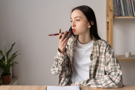 Beautiful caucasian teenage girl giving voice commands to virtual assistant on smartphone, talking while recording audio messages, using mobile phone, smiling. Hand with mobile phone close upの写真素材