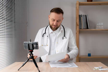 Caucasian male doctor in a lab coat holds documents in his hands and talks with colleagues during a video call on a smartphone.の写真素材