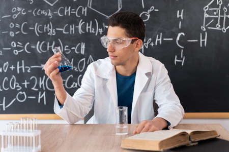 A young Arab male chemistry teacher sits at a table against the background of a chalk board with different formulas, dressed in a white coat and goggles, holding a test tube in a blue liquid.の写真素材